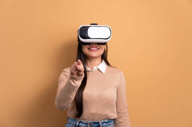 Neşeli genç bir kadın, VR kulaklığı kullanıyor ve bej rengi stüdyo arka planında el kol hareketi yapıyor. sanal gerçeklik, teknoloji simülasyon kavramı.
