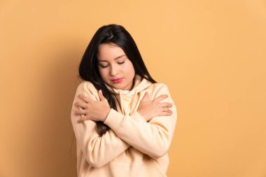 Beyaz tenli, kendini kucaklayan, bej renkli stüdyo geçmişiyle sarılan bir kadın. Kendini sevme, bakım konsepti.