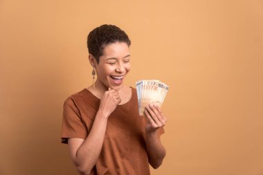 Brezilya parasına bej renklerle bakan mutlu siyahi genç bir kadın. Ekonomi, kredi, ödeme, başarılı konsept.