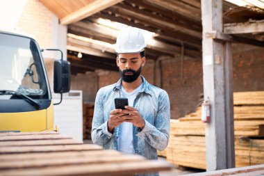 Professional contractor at work. Confident Successful african american man with hard hat texting and typing on the mobile smart phone in construction site.