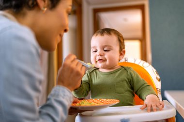 Yeşil elbiseli neşeli çocuk, sevgi dolu anne tarafından kaşıkla besliliyor. Sevimli çocuk evdeki yüksek sandalyede sağlıklı bir yemeğin tadını çıkarıyor..