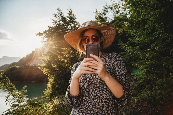 Güneş gözlüklü ve şapkalı bir kadın göl kenarında cep telefonu kullanıyor.