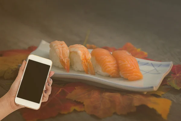 Mujer mano mantenga el teléfono inteligente en borrosa conjunto de sushi japonés — Foto de Stock