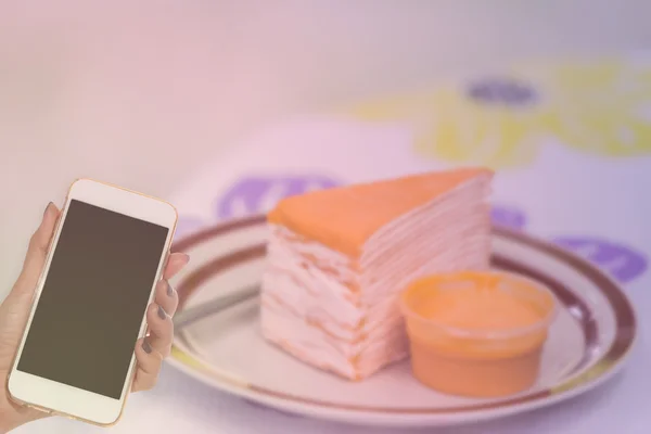 Mujer mano mantenga el teléfono inteligente en pastel de naranja borrosa — Foto de Stock