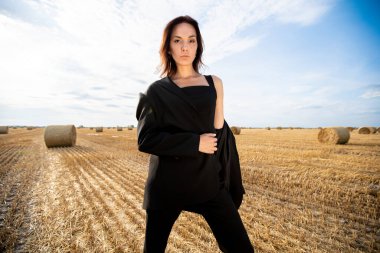 Yazın siyah takım elbiseli genç bir kız sıcak bir günde samanlıklarla dolu bir tarlada duruyor.