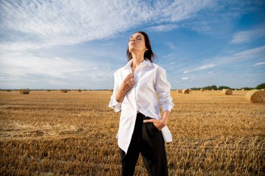  Yazın beyaz gömlekli genç bir kız sıcak bir günde samanlıklarla dolu bir tarlada duruyor.