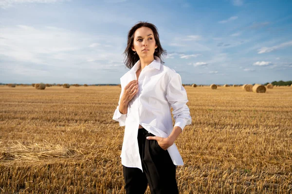  Yazın beyaz gömlekli genç bir kız sıcak bir günde samanlıklarla dolu bir tarlada duruyor.