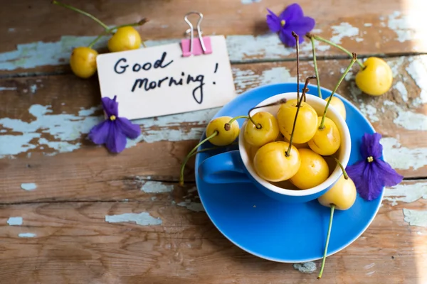 Blaue Teetasse voll mit gelben Kirschen mit Veilchen in einem Topf — Stockfoto