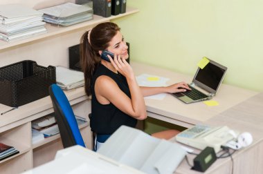 Young businesswoman sitting at desk, talking on cell phone.