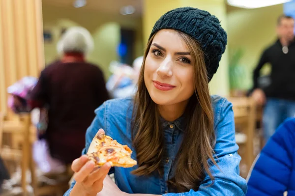 A young woman sitting in a restaurant, smiling,looking at the camera holding a slice of pizza. — Zdjęcie stockowe