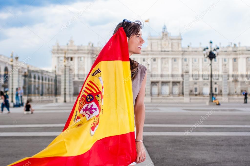 Young woman in front of Palacio de Oriente - the Royal Palace of Madrid, holding a flag with a big smile.