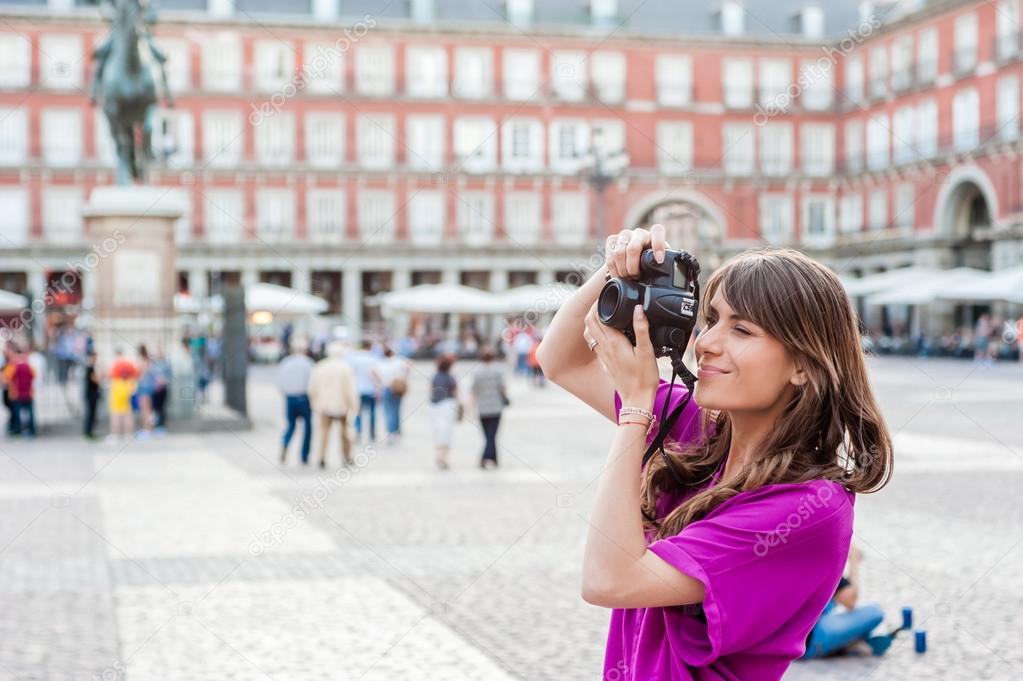 Young woman tourist holding a photo camera, sitting and looking at buildings in Plaza Mayor square, Madrid, Spain