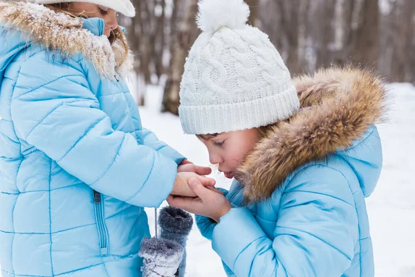 Little girl holds sister\'s cold hands, breathes, warms them. Walking, having fun in snowy winter park. Blue jackets with fur, hat, mittens. Family picnic in cold weather. Outdoor activity.