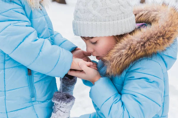 Little girl holds sister\'s cold hands, breathes, warms them. Walking, having fun in snowy winter park. Blue jackets with fur, hat, mittens. Family picnic in cold weather. Outdoor activity.