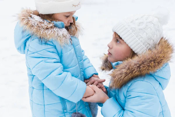 Little girl holds sister\'s cold hands, breathes, warms them. Walking, having fun in snowy winter park. Blue jackets with fur, hat, mittens. Family picnic in cold weather. Outdoor activity.