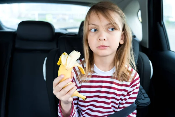 Little blond caucasian girl is driving in car, eating banana. Traveling on road in safe baby seats with child belts. Fun family trip, activity with parents.