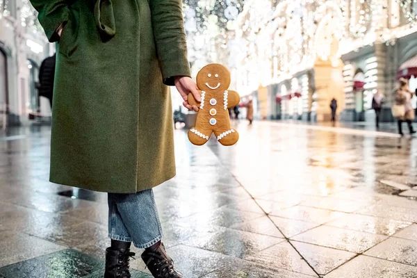 Young beautiful girl, woman with huge funny gingerbread man. Christmas shopping. Walking on market street in big city decorated with garlands, lights.Stylish look, wool coat, hat.New year celebration.