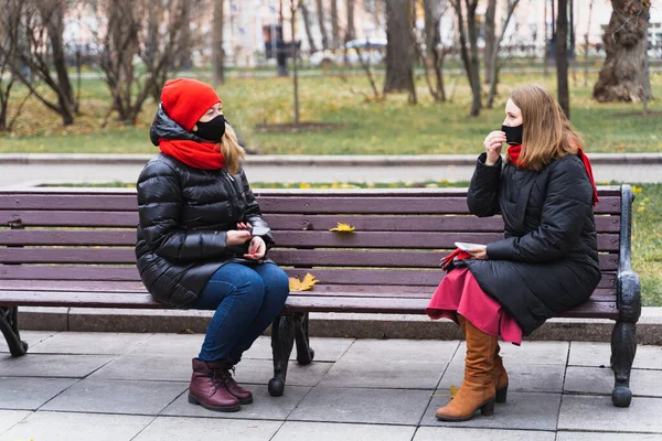 Two young women, friends in face masks using, speaking mobile phone on safe social distance. Sitting on bench in autumn park.Meeting,talking during Covid-19 coronavirus pandemic.Warm jacket,red scarf.