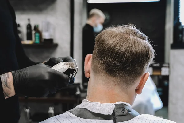 Visit to the barbershop. Hairdresser, barber shaves client\'s temples,sides with hair clipper. Young stylish man makes fashionable haircut.Customer sitting in dark men\'s beauty salon covered with cape.