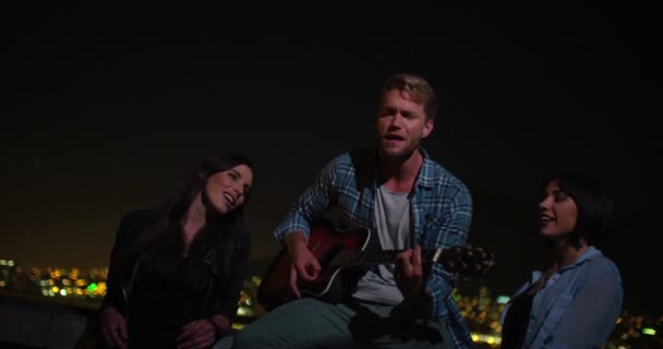 Мужчина играет на гитаре и поет с девушками на вечеринке на крыше — стоковое видео