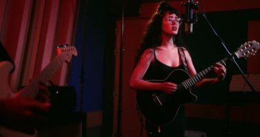 Sanatçı müzik stüdyosunda gitaristle şarkı söylüyor ve gitar çalıyor.