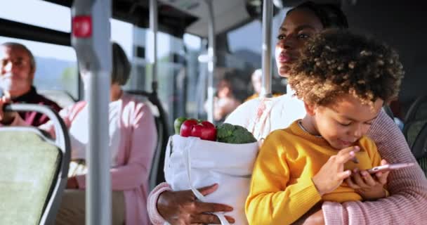 Afroamerykańska matka trzyma zakupy, syn bawi się na smartfonie w autobusie — Wideo stockowe