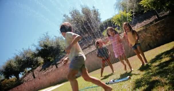 เด็ก ๆ เล่นและกระโดดข้ามสปริงเกอร์ในสวน — วีดีโอสต็อก