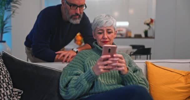 Старша жінка, що показує смартфон чоловікові сидячи на дивані — стокове відео