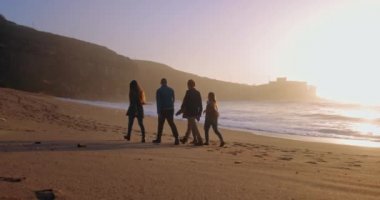 Arkadaş grubu silueti gün batımında sahilde yürüyor.