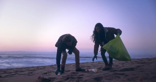 Два друга собирают мусор на пляже на закате — стоковое видео