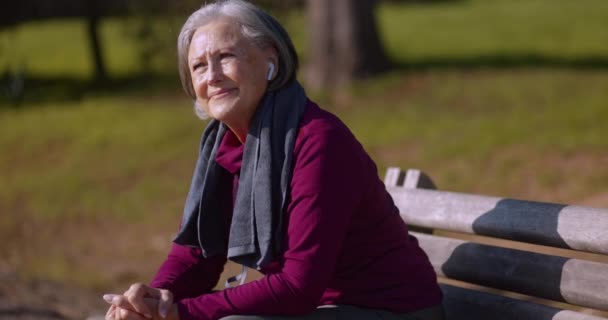 Seniorin mit Kopfhörern sitzt nach Übung auf Bank — Stockvideo