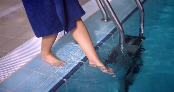 Woman in bathrobe dipping foot into swimming indoor pool — 비디오
