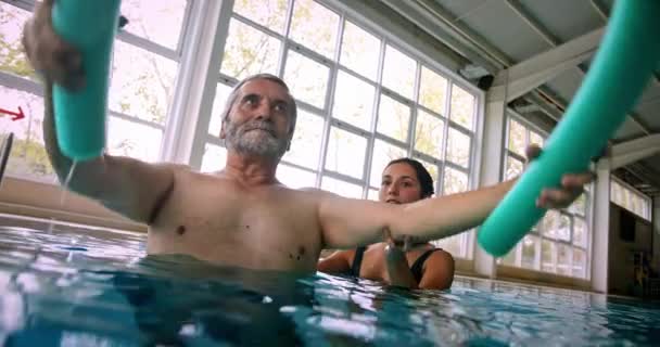 Instrutor ajudando o homem sênior levantando macarrão piscina na aula de aeróbica — Vídeo de Stock