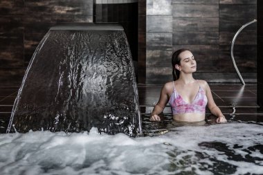 Bikinili genç ve güzel bir bayan şelaleli yüzme havuzunun köpüklü suyunda oturuyor ve spa kulübünde hidroterapi seansının keyfini çıkarıyor.