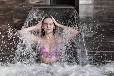 Pembe bikinili çekici genç bir kadın şelale jeti suyunun altında duruyor ve kaplıca merkezinde hidroterapi prosedürünün tadını çıkarıyor.