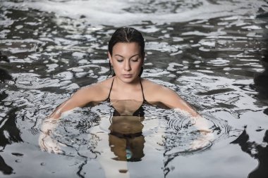 Yüksek açılı dingin genç esmer havuzda taze kristal suyun tadını çıkarırken kaplıca merkezinde hidroterapi prosedürü uygulanıyor.