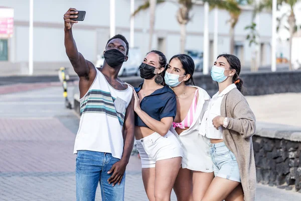 Group of smiling multiracial friends wearing masks standing in city and taking selfie on smartphone while spending time together in summer on Lanzarote