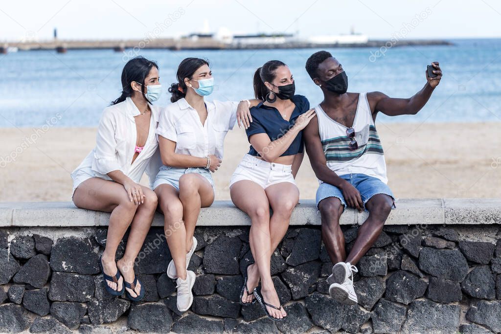 Group of diverse friends in masks sitting on stone border near beach and taking selfie on smartphone while enjoying weekend on Lanzarote