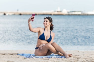 İspanya 'nın Lanzarote adasında deniz kenarında dinlenirken bikinili, gülümseyen ve selfie çeken hoş bir kadın.