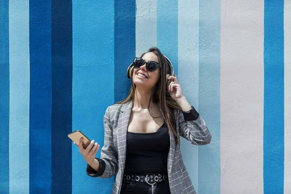 スマートフォンを持つ幸せな若い女性は笑顔で 街の通りの縞模様の壁に対してヘッドフォンでお気に入りの音楽を聞きながら検索 — ストック写真