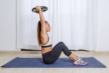 Spor giyimli, etkin, genç ve güçlü bir bayanın tüm vücut manzarası. Paspasın üzerinde oturup ağırlık tabağını kaldırarak evde tek başına spor yaparken.