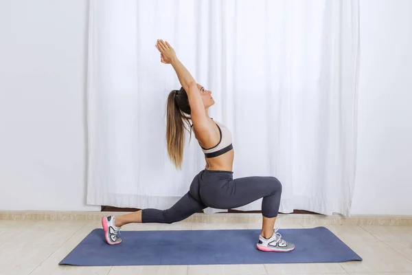 Full body side view of fit female in sportswear and sneakers standing in Crescent lunge position with arms raised while stretching body during fitness yoga training at home