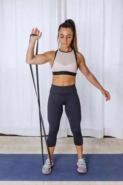 Full body of young fit female in sportswear doing one arm bicep curl exercise with resistance rope during fitness workout at home