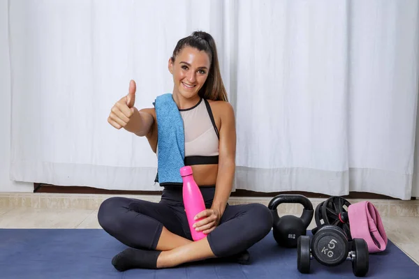 Full body of happy active young female in sportswear showing thumb up gesture while practicing healthy lifestyle and resting after fitness workout with sports equipment at home