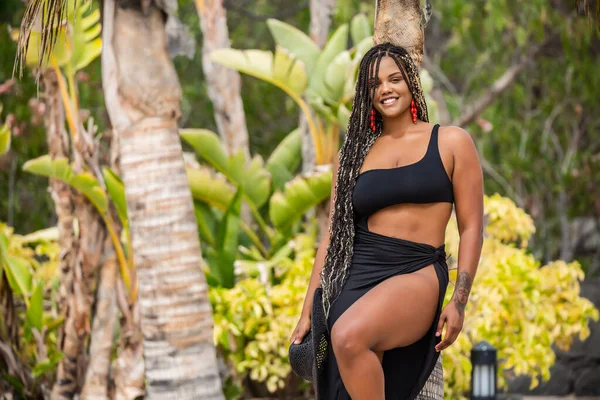 Siyah yaz kıyafetleri içinde, palmiye ağacına yaslanmış siyah şapkalı, gülümseyen güzel bir kadın. Elinde siyah şapkası ve siyah yaz kıyafetleri olan Afro-Amerikalı kadın kameraya bakıyor..