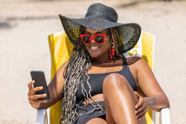 Plajda telefon kullanan siyah şapkalı ve güneş gözlüklü Afro-Amerikalı bir kadın. Neşeli yetişkin kadın sahilde bir yaz gününün tadını çıkarıyor..