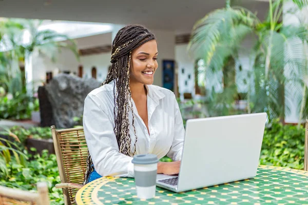Yetişkin Afro-Amerikalı bir kadın dışarıdaki bir kafede dizüstü bilgisayar kullanıyor, bitki ve yeşilin odak noktası dışında. Örgülü bir kadın dizüstü bilgisayar kullanıyor ve kahve içiyor..