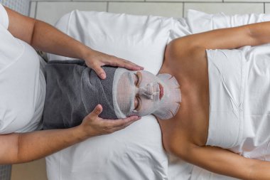 Güzellik uzmanının, kaplıca salonunda cilt bakımı için yüzünü gazlı bezle kaplamış bir kadının kafasına havlu dayadığı ön manzara. Spa kavramı.