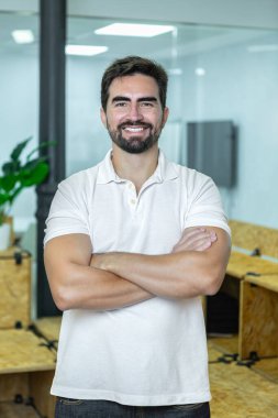 Profesyonel bir adam, kollarını aynı yerde kavuşturarak güvenle ayakta durur. İç tasarım modern tasarım elementleri ve ahşap mobilyalar içerir..