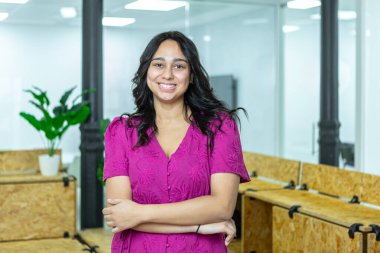 Genç bir kadın, kendine güvenen bir şekilde birlikte çalışma alanında duruyor. Gülümsüyor ve kollarını kavuşturuyor. Çalışma alanında ahşap masalar ve bitkiler yer alıyor..